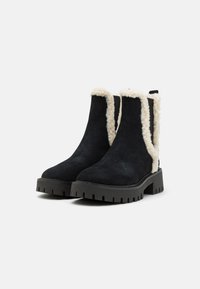 Black suede ankle boots with cream faux fur lining, chunky black tread sole, and minimalist design. Side cut-outs enhance breathability.