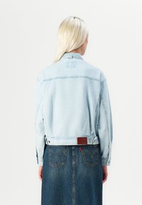 Light blue denim jacket with a cropped design, featuring a leather patch at the back and buttoned cuffs, worn over a dark denim skirt.