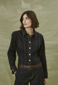 Žena s krátkými hnědými vlasy, oblečená v tmavé džínové bundě a kalhotách se zlatými knoflíky a hnědým opaskem, na texturovaném světle zeleném pozadí.