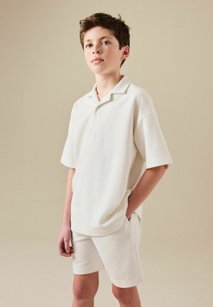 Junger Junge mit kurzen braunen Haaren und Sommersprossen trägt ein übergroßes weißes Polohemd und passende Shorts, steht mit einer Hand in der Tasche.