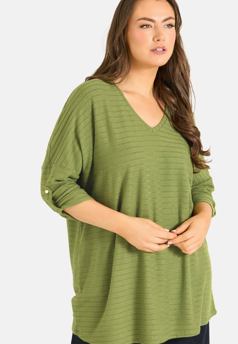 Donna che indossa un maglione verde a coste con scollo a V, maniche risvoltate fissate da un bottone, mani intrecciate in vita, guarda di lato.