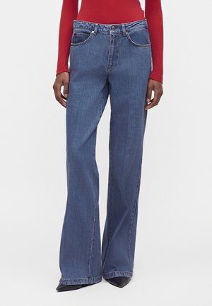 Persoon draagt blauwe wijdere jeans, rood top met lange mouwen en zwarte puntige schoenen, staand tegen een effen lichte achtergrond.