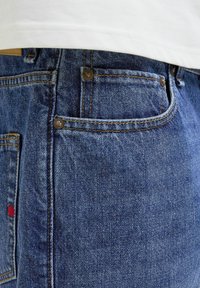 Jeans in denim blu scuro con cuciture a contrasto, dotati di una tasca frontale, rivetti in rame e un piccolo etichetta rossa sulla tasca posteriore.