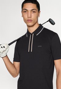 Polo noir à manches courtes avec col zippé et rayures d'accent blanc et orange. Porté avec un club de golf et un gant.