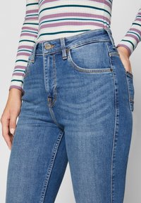 Los pantalones vaqueros de mezclilla azul presentan una cintura alta, cinco bolsillos y costuras naranjas en contraste. El material tiene una textura suave con un ligero estiramiento.