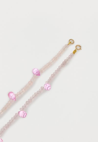 Deux bracelets roses en perles avec des pierres roses rondes et en forme de larme, ainsi que des fermoirs circulaires de couleur or sur fond blanc.