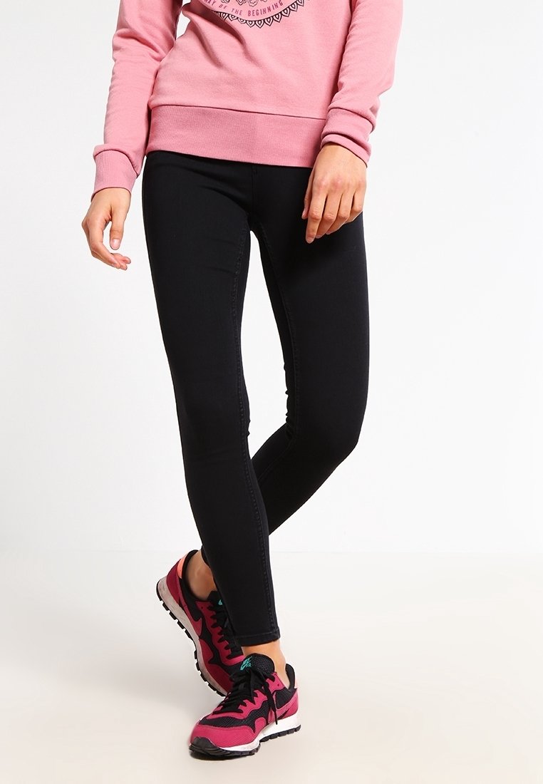 Jean slim noir en denim, avec une texture lisse. Porté avec des chaussures de sport rose et noir. Le haut est rose pastel.