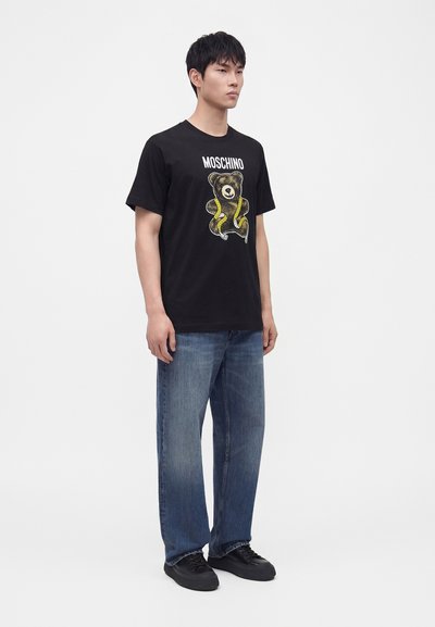 Νεαρός άνδρας που φοράει μαύρο T-shirt Moschino με γραφικό αρκουδάκι, μπλε τζιν και μαύρα αθλητικά παπούτσια, στέκεται μπροστά σε λευκό φόντο.