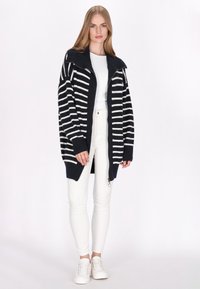 Cárdigan a rayas azul marino y blanco con un amplio cuello, diseño de frente abierto y puños acanalados, combinado con pantalones blancos y zapatillas blancas.
