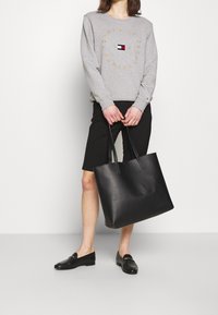 Felpa grigia con logo, pantaloni bermuda neri e borsa in pelle nera con manici lunghi. Modella indossa mocassini neri.