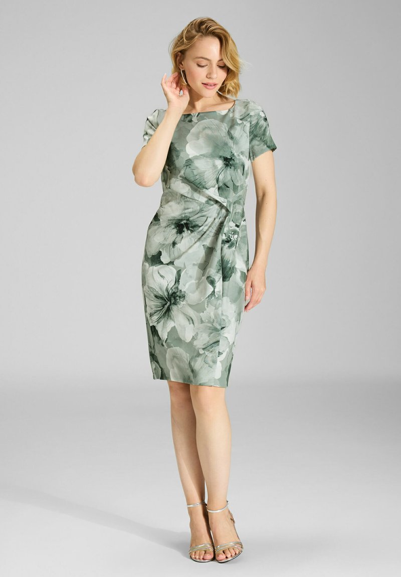 Robe fleurie aux tons verts, manches courtes, longueur genou, avec un design drapé et des motifs floraux proéminents. Associée à des sandales argentées à brides.