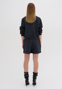 Donna con capelli castano chiaro e lisci, che indossa un top lungo nero, pantaloni corti neri e stivali neri con tacco, in piedi voltata di spalle.