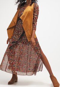 Květinové maxi šaty z průsvitné texturované látky s bočními rozparky, vrstvené s bezrukávovou hnědou semišovou vestou s hroty. Hnědé kotníkové boty.