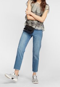 Lätt, genomskinlig topp med ormskinnsmotiv; avslappnade blå jeans med fransade fållar; silverfärgade sneakers med glittrande detaljer. Svart axelremsväska.