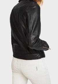 Czarna skórzana kurtka motocyklowa o dopasowanym kroju, z pikowanymi akcentami na plecach i zamkami błyskawicznymi przy mankietach, w połączeniu z białymi dżinsami.