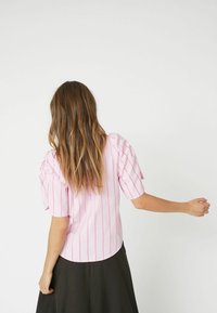 Blusa rosa chiaro con righe bianche verticali, maniche corte a sbuffo e orlo arrotondato, con dettagli testurizzati sulle spalle delle maniche.