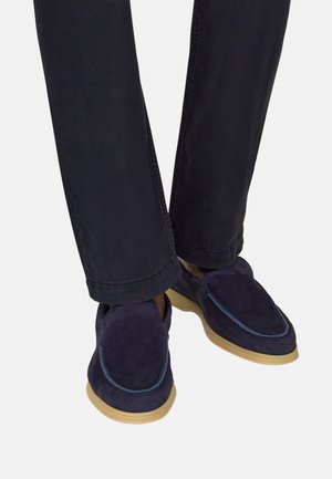 Donkerblauwe suède loafers met beige rubberen zolen, gedragen met donkerblauwe broek tegen een lichte achtergrond.