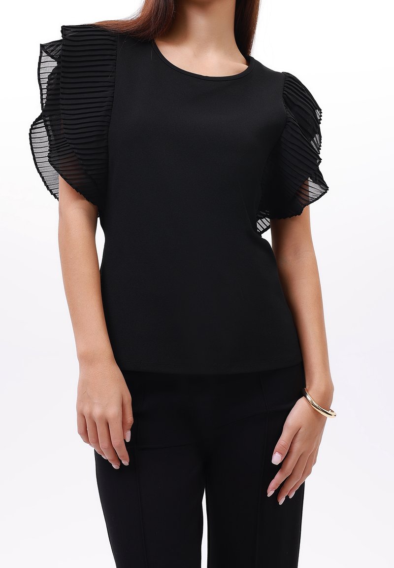 Czarna bluzka z krótkimi, plisowanymi, przejrzystymi rękawami. Gładki materiał, okrągły dekolt, dopasowany krój. Nosi się ją z czarnymi spodniami i złotą bransoletką.