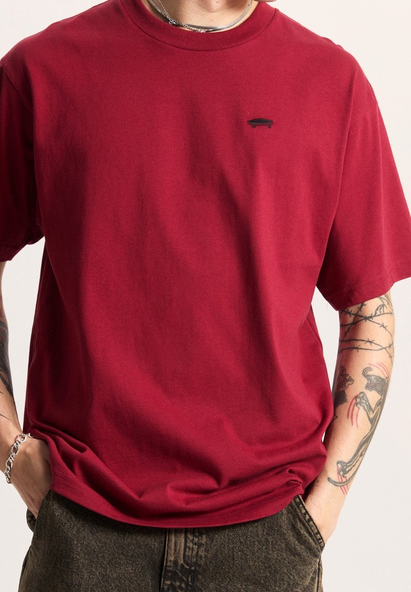 Personne portant un t-shirt rouge avec un petit logo skateboard noir, bras droit tatoué, les mains dans les poches d'un pantalon sombre.