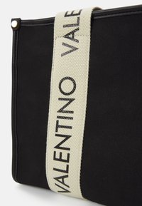 Schwarze Stofftasche mit strukturierter Oberfläche, ausgestattet mit einem breiten cremefarbenen Tragegurt, der mit schwarzer Schrift "VALENTINO" beschriftet ist. Meschenfarbene Hardware-Details.