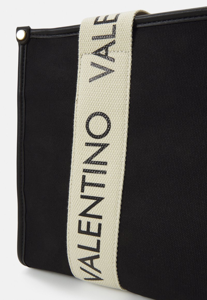 Bolso de tela negra con una superficie texturizada, que presenta una correa ancha crema etiquetada como "VALENTINO" en letras negras. Detalle de herrajes en color dorado.