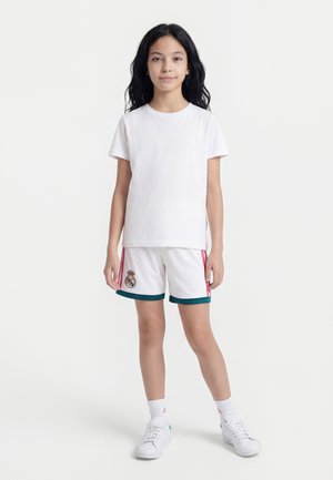 Niña joven con cabello largo y oscuro está de pie vistiendo una camiseta blanca y pantalones cortos de fútbol blanco del Real Madrid con zapatillas blancas sobre un fondo liso.