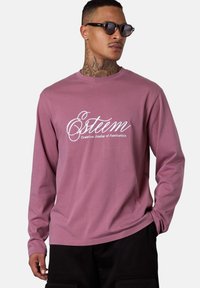 Mann mit Nackentattoos und Sonnenbrille, der ein mauvefarbenes Langarmshirt mit dem Text "Esteem Creative Atelier of Aesthetics" trägt, mit entspannten Händen.