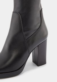 Bottine en cuir noir avec un bout rond, une texture lisse et un talon bloc épais. Comprend des coutures ton sur ton et un panneau élastique latéral.