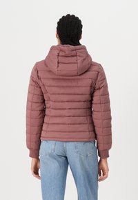Wypełniona, bordowa kurtka z kapturem, z poziomym pikowaniem, ściągaczami przy mankietach i dopasowanym krojem. Noszona z jasnoniebieskimi dżinsami.