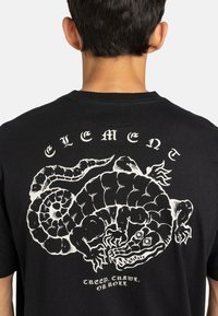 Zwarte katoenen t-shirt met een grote witte afbeelding van een slang met ingewikkelde details en het woord "ELEMENT" erboven.