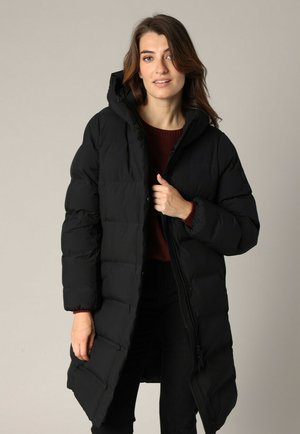 Manteau d'hiver noir avec une capuche, présentant un design matelassé, des fermetures éclair et des poches latérales, porté sur un pull bordeaux.