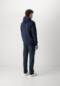 Felpa blu navy con cappuccio attaccato, polsini e orlo a costine. Indossata con jeans scuri e sneakers bianche. Lo sfondo è semplice e chiaro.