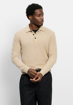Homme portant un pull beige en maille à manches longues avec des boutons noirs, debout, les mains jointes, regardant sur le côté devant un fond uni.