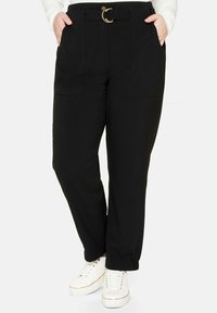 Osoba v černých kalhotách s vysokým pasem, opaskem a kapsami, v kombinaci s bílými teniskami a bílým svetrem s dlouhým rukávem.