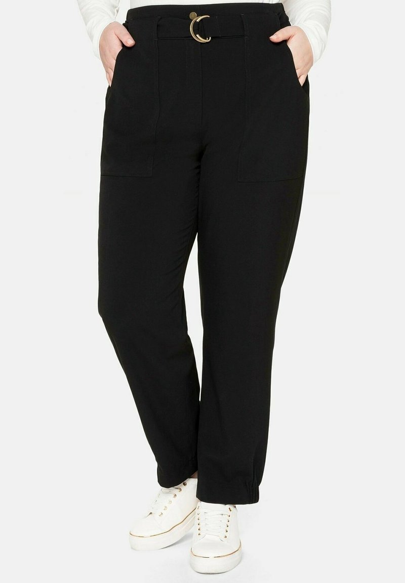 Osoba v černých kalhotách s vysokým pasem, opaskem a kapsami, v kombinaci s bílými teniskami a bílým svetrem s dlouhým rukávem.