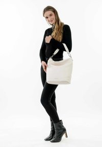 Sac à main en cuir blanc avec une finition texturée, doté d'une poignée supérieure, de quincaillerie en argent et d'un charm circulaire. Stylé avec un pantalon noir et des bottes à talons.