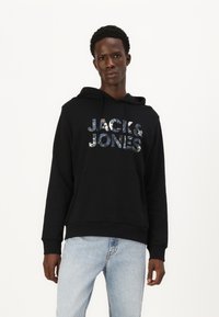 Felpa nera con cappuccio e tasca a marsupio frontale, con logo riflettente "JACK & JONES", indossata con jeans azzurri chiari.