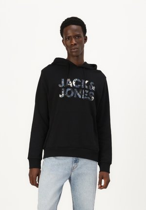 Schwarzer Kapuzenpullover mit vorderer Kängurutasche, versehen mit einem reflektierenden "JACK & JONES" Logo, getragen mit hellblauen Jeans.