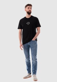 Camiseta de algodón negra con un diseño de logo en el pecho, combinada con jeans de mezclilla azul claro y zapatillas grises con toques en blanco y marrón.