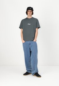Tricou gri din bumbac cu logo pe piept, blugi deschiși din denim, cu croială lejeră, și pantofi sport negri. O șapcă neagră completează ținuta.