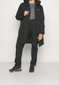 Chaqueta negra impermeable con capucha, que cuenta con bolsillos frontales y una cremallera, combinada con pantalones negros y zapatos de senderismo negros.