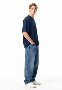 Temno modra oversize majica s kratkimi rokavi, s svetloblue širokimi kavbojkami in belimi supergami, stoji ob enostavni beli podlagi.