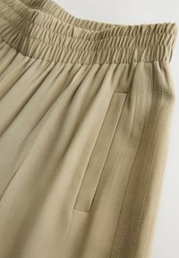 Beige, plissee bukser med elastisk talje og sidelommer. Stoffet ser glat ud med synlige syningsdetaljer.