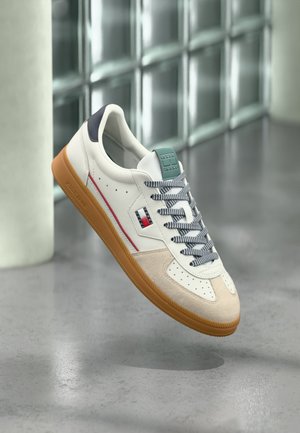 Adidași albi și bej cu talpă de gumă, șireturi alb-negru, logo lateral roșu și albastru, plutind deasupra podelei gri din beton lângă un perete din blocuri de sticlă.
