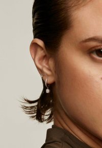 Pendientes de aro de plata con un diseño texturizado y un pequeño charm en forma de diamante colgante. También es visible un pendiente de botón en la oreja.