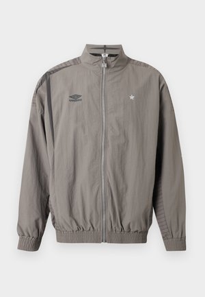 Light gray zip-up sports jacket with Umbro logo on left chest and star emblem on right chest, elastic cuffs and hem, and striped shoulder detail.