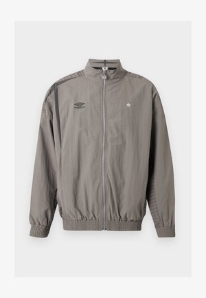 Chaqueta deportiva gris claro con cremallera, logo de Umbro en el pecho izquierdo y emblema de estrella en el pecho derecho, puños y bajo elásticos, y detalle de rayas en los hombros.