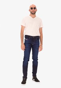 Homme chauve avec une barbe portant des lunettes de soleil rondes, un t-shirt blanc en V, un jean bleu foncé, des baskets noires et un bracelet, se tenant sur un fond blanc.