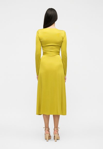 Frau mit langen dunklen Haaren, die ein senfgelbes Midi-Kleid mit langen Ärmeln und beigen High-Heel-Sandalen trägt, steht mit dem Rücken zur Kamera.