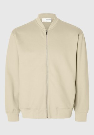 Veste bomber beige en tissu doux, présentant une fermeture éclair, un col et des manches côtelés, ainsi que des poches latérales. Design lisse et minimaliste.